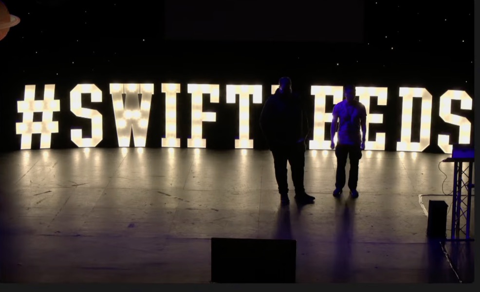 Shadowy outlines of me and James as we stand on the dark stage with #SwiftLeeds is spelled out brightly behind us.