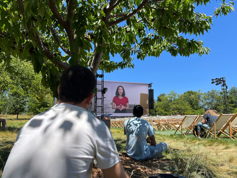 Sitting under a tree watching a large screen. Novall Swift is on the screen.